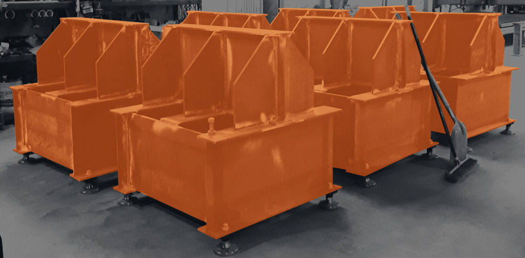 Several large, orange-painted metal industrial parts, ready for custom load testing, are arranged in rows on a workshop floor next to a broom.